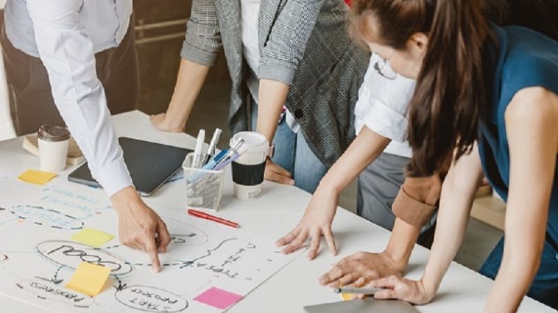 Drei Personen beugen sich über einen Tisch und zeigen auf Pläne