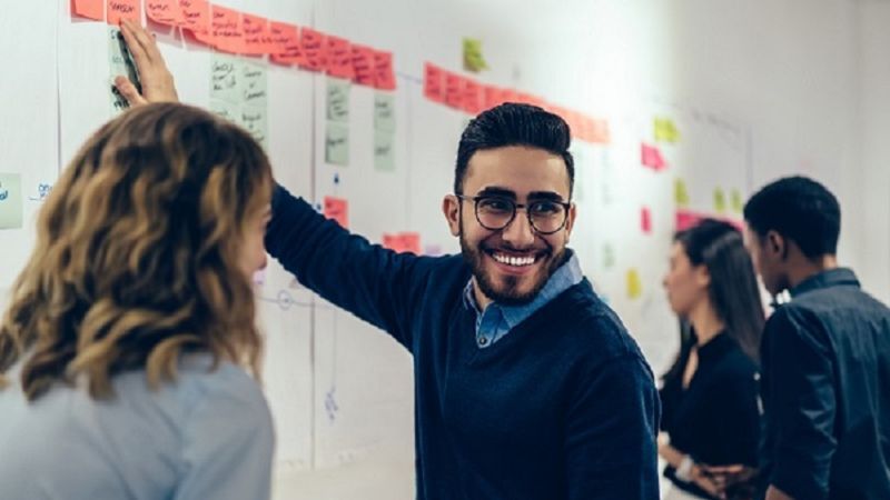 Ein junger Mann lächelt und zeigt auf eine Wand gefüllt mit Notizen